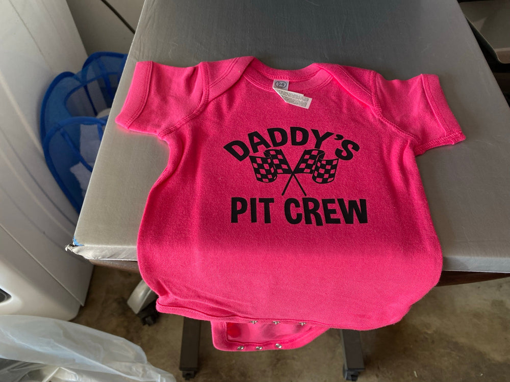 Bright pink baby bodysuit with black Daddy’s Pit Crew lettering and crossed checkered racing flags, printed by Minty Tees.