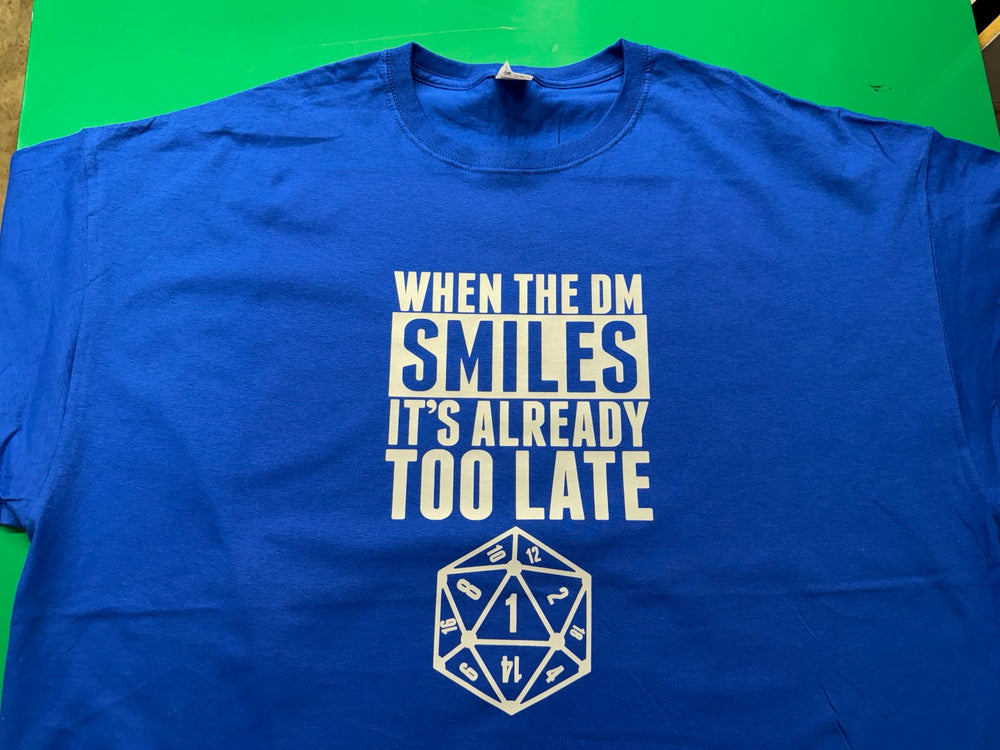 Royal blue T-shirt with pale lettering reading When the DM Smiles It's Already Too Late and a twenty-sided die front print, printed by Minty Tees.