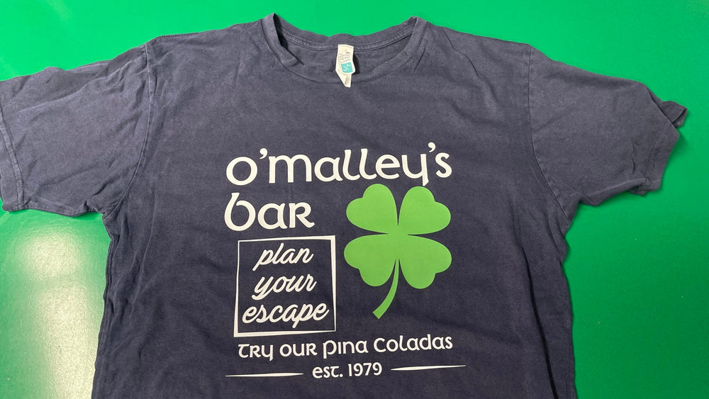 Dark gray O’Malley’s Bar fan tribute T-shirt with cream lettering and a bright green four-leaf clover.