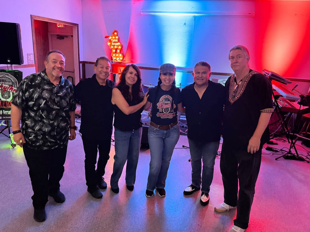 Group photo at a Rhythm Jukeboxx performance, with a woman wearing a Rhythm Jukeboxx T-shirt.
